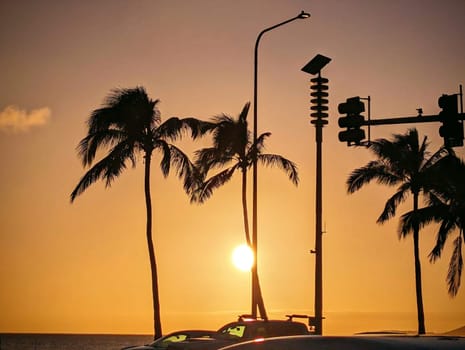 A mesmerizing view of the sunset off Oahu, Hawaii, where the golden sun dips near the horizon, casting a warm glow that silhouettes palm trees and street lights, embodying the tranquil and enchanting evenings of the island. 