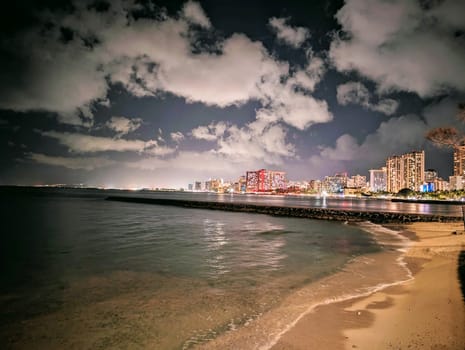 A captivating view of Waikiki at night, where the towering buildings meet the serene beach, and the rhythmic waves of the ocean create a harmonious blend of urban development and natural beauty. The shimmering waters reflect the vibrant city lights, inviting visitors to explore this iconic Hawaiian neighborhood. 