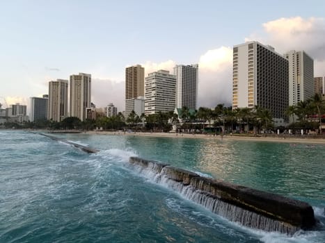 A captivating view of Waikiki, where the towering buildings meet the serene beach, and the rhythmic waves of the ocean create a harmonious blend of urban development and natural beauty. The shimmering waters reflect the vibrant city lights, inviting visitors to explore this iconic Hawaiian neighborhood.