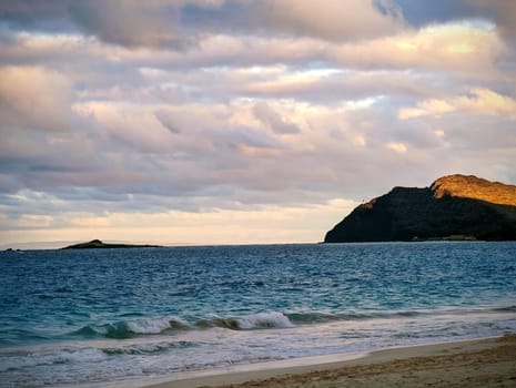 Capture the tranquil beauty of Waimanalo Beach at sunset, where the gentle waves kiss the pristine sands. In the distance, a rock island and lighthouse stand as silent witnesses to the dance of nature, while the setting sun casts a golden hue over the clouds and waters.