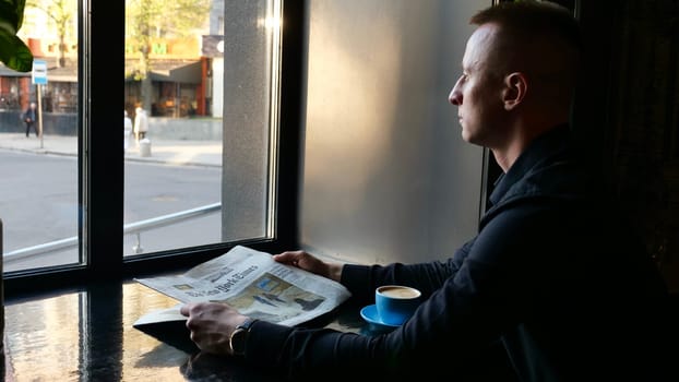 A young seriously student in a business suit holds and reading a paper newspaper the new york times inside in a cafe