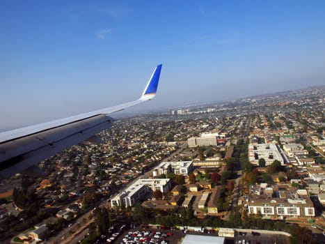 LA - October 26, 2018: An awe-inspiring aerial view captures the sprawling cityscape of Los Angeles as seen from an airplane during approach. The wing of the aircraft, painted in shades of gray with a blue and white tip, frames the scene. Below, the cityscape unfolds with numerous buildings, roads, and structures characteristic of a densely populated area. The sky, mostly clear with scattered clouds, provides a serene backdrop. A faint haze envelops the city, perhaps a result of atmospheric conditions or urban activity.