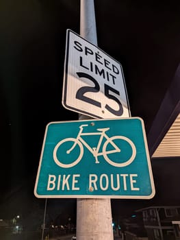 Honolulu - March 21, 2023: Two essential road signs illuminated under the city lights at night. The top sign indicates a speed limit of 25, ensuring drivers maintain a safe speed, while the vibrant green sign below marks the path as a designated bike route, highlighting the city’s commitment to safety and eco-friendly transportation options.
