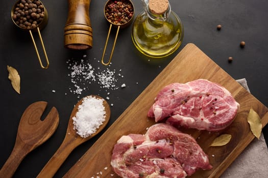 Two raw pork neck steaks on a board and spices for cooking. Top view of black table. 