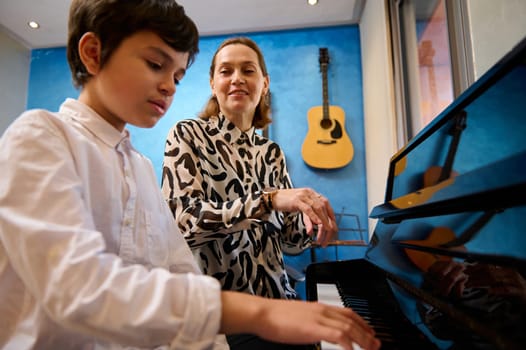 Caucasian teenager pianist playing piano under the guidance of his music teacher at home. Handsome adolescent boy performing classical melody on grand piano. Acoustic guitar hanging on a blue wall