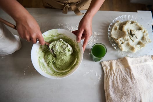 Directly above woman's hands using a wooden spoon mixing ingredients and kneading dough for making homemade ravioli or dumplings stuffed with mashed potatoes, standing t kitchen table with ingredients