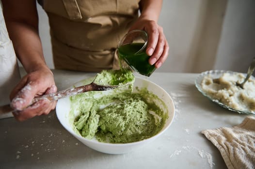 Close-up hands of a chef pastry, housewife pouring green spinach water into a bowl with wheat four, making dough of green color for ravioli or dumplings with mashed potato, in the rustic home kitchen