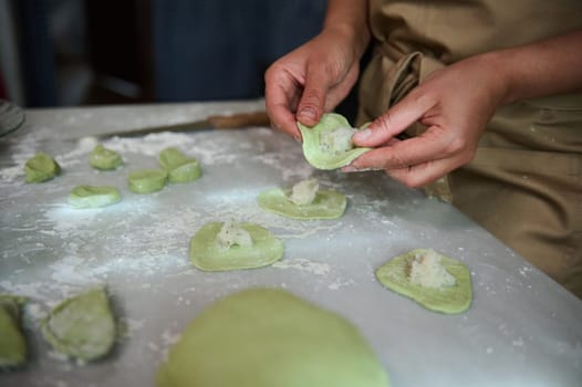 Close-up woman's hands stuffing dough with mashed potatoes, modeling dumplings, standing at kitchen table with kneaded dough and ingredients. The process of cooking ravioli, varenniki, vegan pelmeni