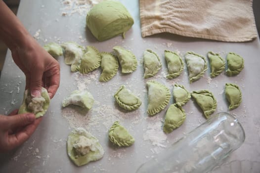 Details on the hands of a woman housewife, chef in beige apron making dumplings from green dough with melted spinach, and filling it with mashed potatoes, standing at kitchen table in rural house