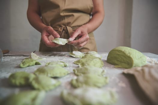 Housewife filling rolled dough with mashed potatoes, preparing homemade varenyky, dumplings, ravioli according to traditional family recipe, standing at marble kitchen table with ingredients. Top view