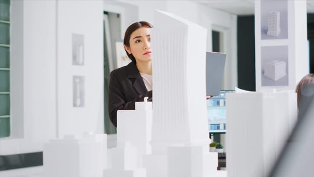 Developer working with laptop to gather scale elements, looking at 3d printed model of building maquette. Technician reviewing creative architectural plan for new construction project.