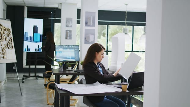 Architectural technician examines 3d building model in creative agency coworking space, taking notes of design lines and measurements. Woman measuring maquette scale and elements. Tripod shot.