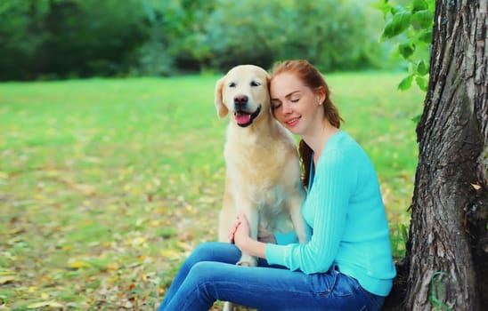 Happy owner young woman with Golden Retriever dog sitting together under tree in summer park