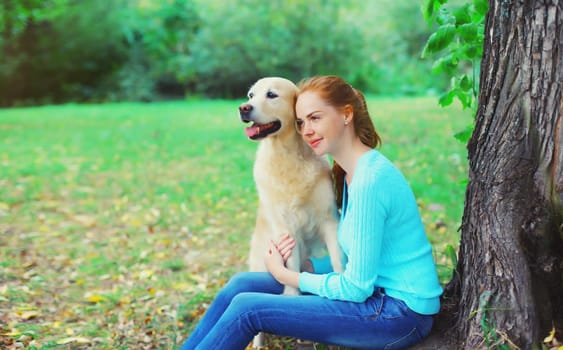 Happy owner young woman with Golden Retriever dog sitting together under tree in summer park