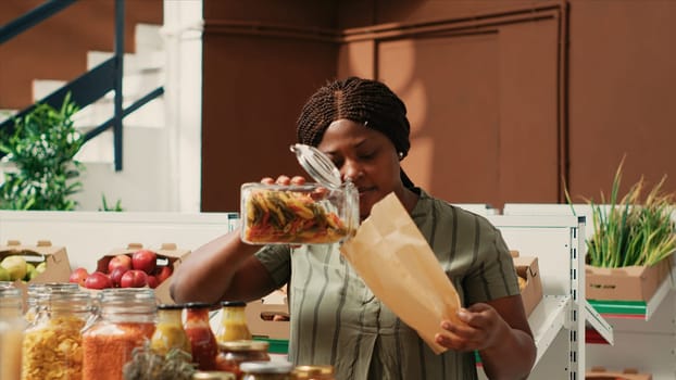 Woman client filling in paper bag with pasta sold as bulk item, buying various types of organic additives free products from local farmers market. Buyer searching for bio pantry supplies.