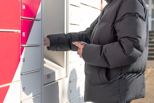 One young Caucasian unrecognizable man in a dark jacket is holding a mobile phone in his hand, and the other is entering information on the screen to receive his parcel from the box, standing on the street on a spring day, side view close-up.