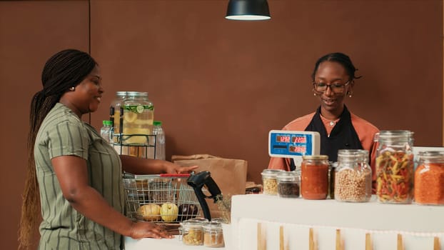 Shopper bringing her natural groceries selection at checkout, talking to vendor about vegan lifestyle and healthy nutrition. Woman searching for ethically sourced farming products.