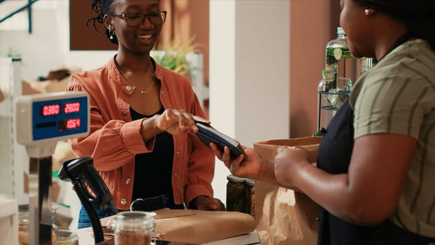 African american client paying for bio goods at checkout, buying freshly harvested ethically sourced produce from local farmers market. Buyer using card at pos terminal, eco friendly.