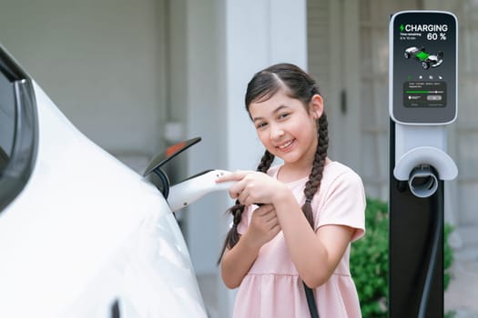 Happy little young girl learn about eco-friendly and energy sustainability as she recharge electric vehicle from home EV charging station. EV car and sustainable future generation concept. Synchronos