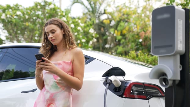 Young woman use smartphone to pay for electricity at public EV car charging station in nature. Modern environmental and sustainable automobile transportation lifestyle with EV vehicle. Synchronos