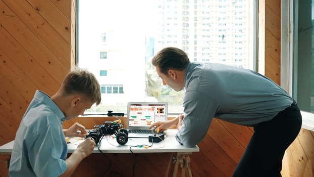 Teacher help student fixing car model and giving advise for programing system or coding prompt while highschool boy listen and use equipment to repair robot at STEM classroom. Back view. Edification.