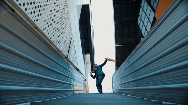 Low angle view of young smiling business man dance between building at center point. Skilled executive manager wearing suit and suitcase and moving to music. Happy investor moving to music. Exultant.