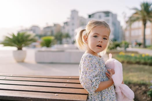 Little girl with a pink soft toy hare stands near a wooden table on the embankment. High quality photo