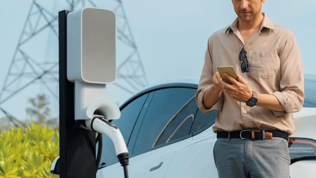 Man pay for electricity with smartphone while recharge EV car battery at charging station connected to power grid tower electrical as electrical industry for eco friendly car utilization.Expedient