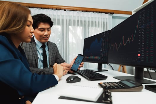 Smiling businessman showing dynamic stock market data on mobile phone to woman writing to memo for analyzing profit value currency rate online website program application at home office. Infobahn.