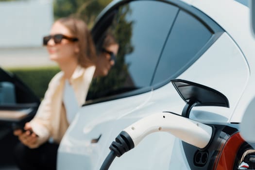 Young woman use smartphone to pay for electricity at public EV car charging station green city park. Modern environmental and sustainable urban lifestyle with EV vehicle. Expedient