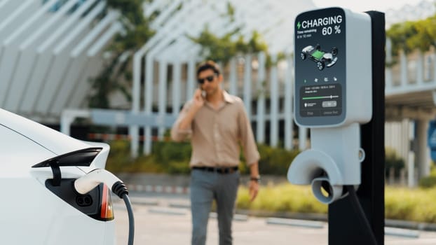 Young man recharge EV electric vehicle at green city mall parking lot while talking on phone. Sustainable urban lifestyle for eco friendly EV car with battery charging station. Panorama Expedient