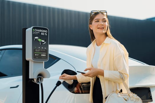 Young woman holding shopping bag and use smartphone to pay for electricity for recharging EV car battery from charging station at city mall parking lot. Modern woman go shopping by eco car. Expedient