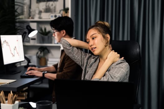 Businesswoman working on desk with stretching arm up and down manner with body health ache of strain overwork while coworker trading stock market on website on pc at late over night time. Infobahn.