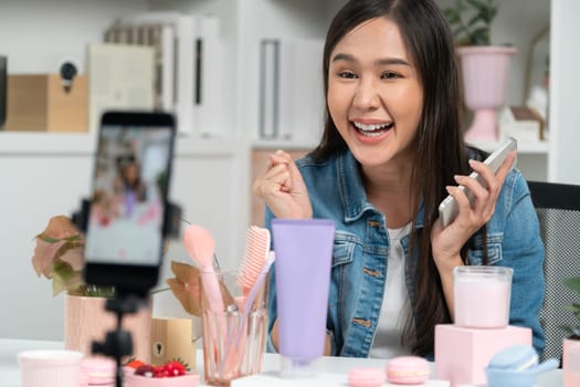 Young beautiful Asian reviewing cosmetic makeup and skin care collection, talking customer on social media online, presenting product surrounded beauty merchandise recording blurred phone . Stratagem.