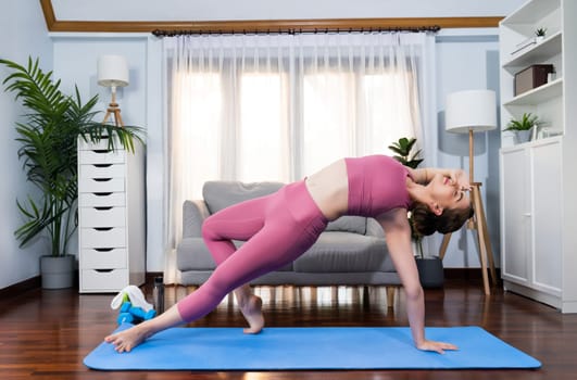 Flexible and dexterity woman in sportswear doing yoga position in meditation posture on exercising mat at home. Healthy gaiety home yoga lifestyle with peaceful mind and serenity.