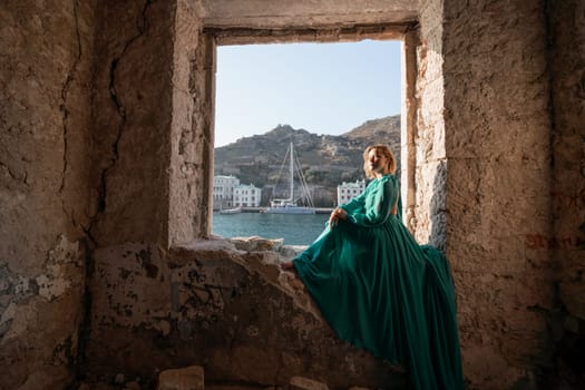 Rear view of a happy blonde woman in a long mint dress posing against the backdrop of the sea in an old building with columns. Girl in nature against the blue sky