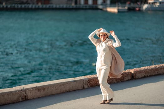 Happy blonde woman in a white suit and hat posing at the camera against the backdrop of the sea.