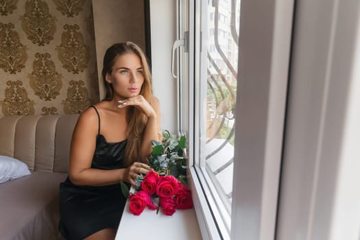 A woman in a black dress is sitting in front of a window with a bouquet of red roses