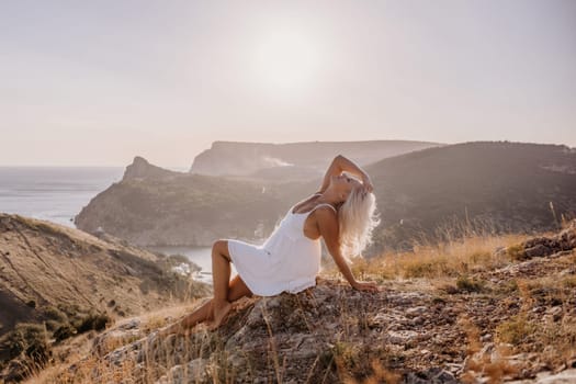 A woman is sitting on a hillside overlooking the ocean. She is wearing a white dress and has blonde hair. The scene is serene and peaceful, with the ocean in the background