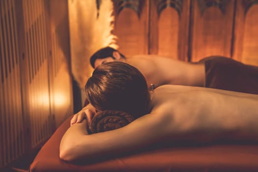 Caucasian couple customer enjoying relaxing anti-stress spa massage and pampering with beauty skin recreation leisure in warm candle lighting ambient salon spa at luxury resort or hotel. Quiescent