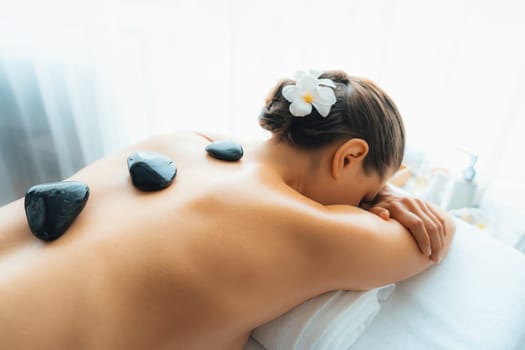 Hot stone massage at spa salon in luxury resort with day light serenity ambient, blissful woman customer enjoying spa basalt stone massage glide over body with soothing warmth. Quiescent