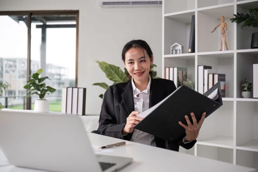 Young Asian business woman in a suit sits and reviews company work documents, checking information on a laptop on a work desk in a white office..
