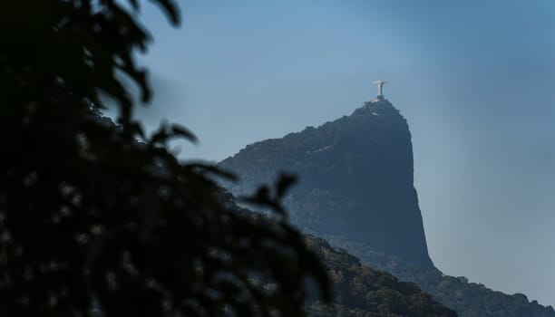 Iconic Christ of Rio de Janeiro seen from afar, atop a mountain amidst lush jungle, against a clear blue sky, with a blurred tree in foreground