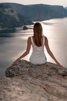A woman sits on a rock overlooking the ocean. She is wearing a white tank top and white shorts. The scene is peaceful and serene, with the woman enjoying the view and the calming sound of the waves