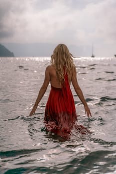 A woman in a red dress is standing in the ocean. The water is calm and the sky is cloudy