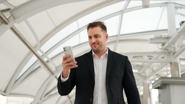 Skilled businessman looking or checking his mobile phone while walking at bridge for going to workplace. Happy manager using telephone to send massage to marketing team and receive good news. Urbane.