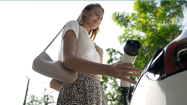 Young woman travel with EV electric car charging in green sustainable city outdoor garden in summer. Urban sustainability lifestyle by green clean rechargeable energy of electric BEV vehicle innards