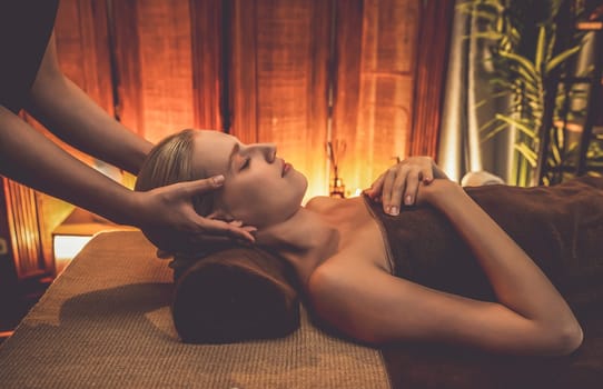 Caucasian woman enjoying relaxing anti-stress head massage and pampering facial beauty skin recreation leisure in warm candle lighting ambient salon spa in luxury resort or hotel. Quiescent