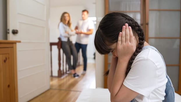 Stressed and unhappy young girl huddle in corner, cover her ears blocking sound of her parent arguing in background. Domestic violence at home and traumatic childhood develop to depression. Synchronos