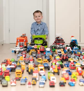 Concept of children's toys. A little boy, 4 years old, cheerful and handsome, happily plays, sitting on the floor, with multi-colored small and large cars in the children's room. Soft focus